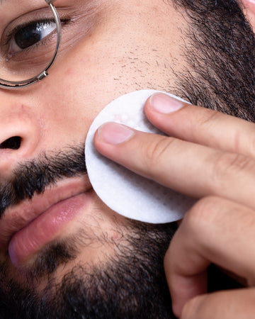 Man with beard and glasses wiping face with cotton pad